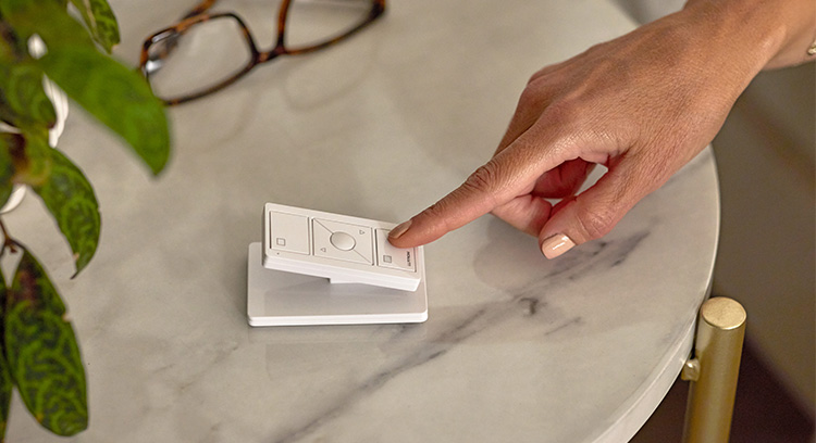 A hand presses a Pico Original Remote for Shades resting on a table next to glasses and a plant.